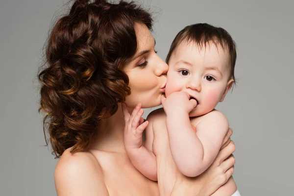 Curly mother with bare shoulders kissing baby boy sucking fingers isolated on grey — Stock Photo