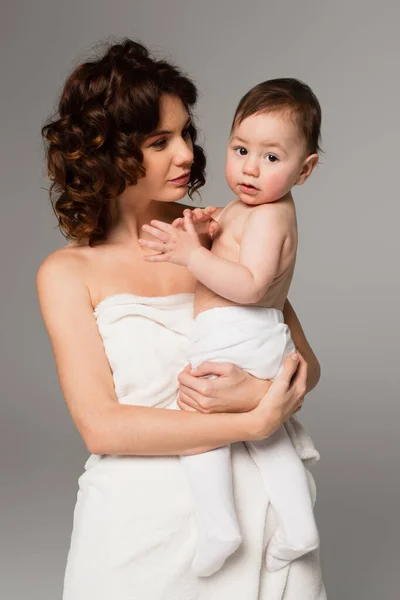 Curly mother wrapped in towel holding baby boy in arms isolated on grey — Stock Photo