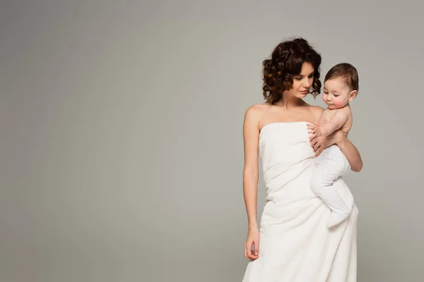 Curly mother wrapped in towel holding baby in arms isolated on grey — Stock Photo