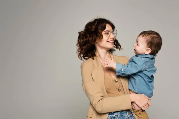 Happy mother in glasses and blazer holding in arms baby boy isolated on grey — Stock Photo