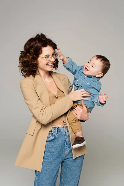 Alegre madre en gafas y chaqueta sosteniendo en brazos feliz bebé niño aislado en gris - foto de stock