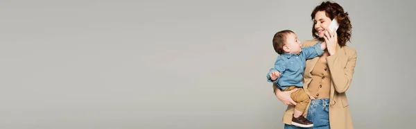 Niño alcanzando teléfono inteligente mientras madre feliz en chaqueta hablando aislado en gris, bandera - foto de stock