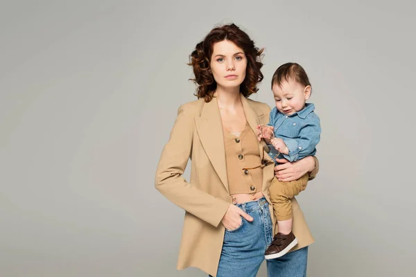 Kleinkind mit Brille, während Mutter im Blazer mit Hand in Tasche auf grau isoliert posiert — Stockfoto