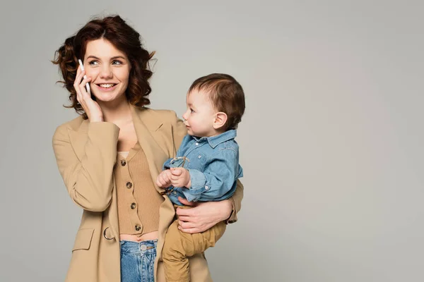 Toddler boy holding glasses while happy mother in blazer talking on smartphone isolated on grey — Stock Photo