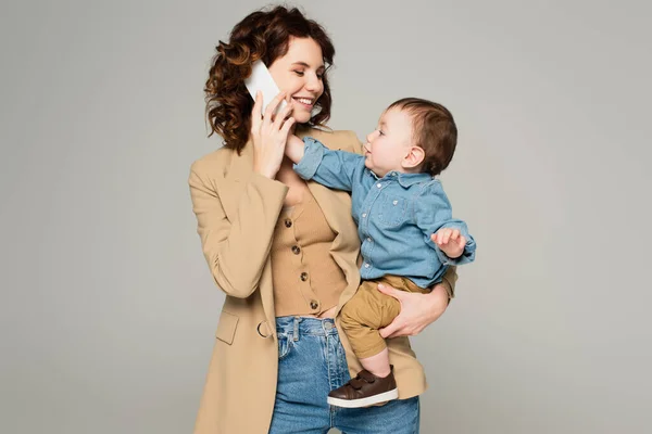 Kleinkind greift zum Smartphone, während glückliche Mutter im Blazer isoliert auf grau spricht — Stockfoto