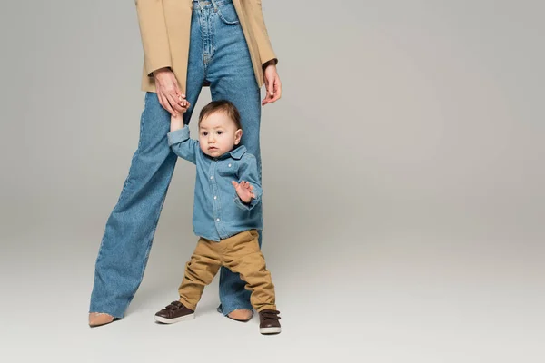 Cropped view of mother supporting and holding hand of baby boy on grey — Stock Photo