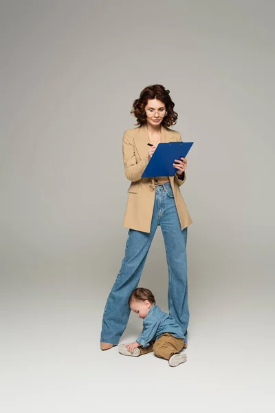 Geschäftsfrau mit Brille schreibt auf Klemmbrett neben Kleinkind Sohn auf grau — Stockfoto