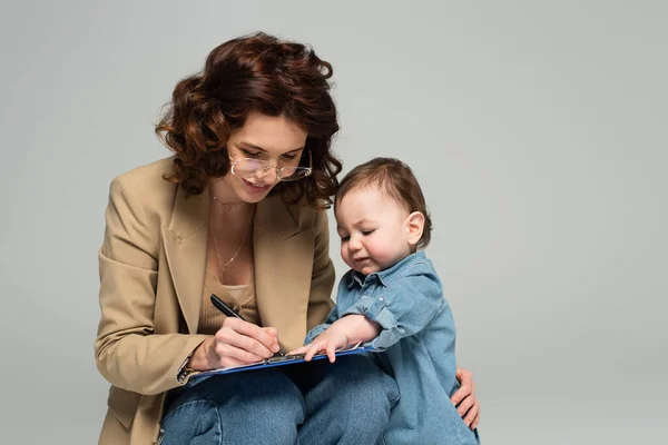 Sonriente mujer de negocios en gafas escribiendo en portapapeles cerca de niño pequeño hijo aislado en gris - foto de stock