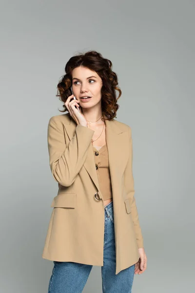 Lockige Frau im beigefarbenen Blazer im Gespräch auf Smartphone isoliert auf grau — Stockfoto