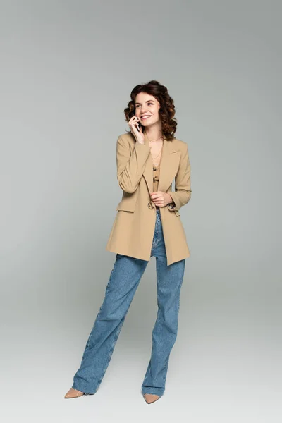 Full length of happy woman in beige blazer talking on smartphone on grey — Stock Photo