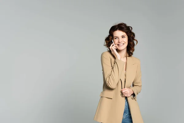 Felice donna in blazer beige che parla su smartphone isolato su grigio — Foto stock