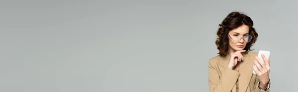 Кучерява жінка в окулярах і бежевий блейзер бере селфі ізольовано на сірому, банер — стокове фото
