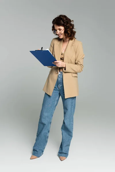 Full length of happy businesswoman in glasses and blazer holding clipboard on grey — Stock Photo