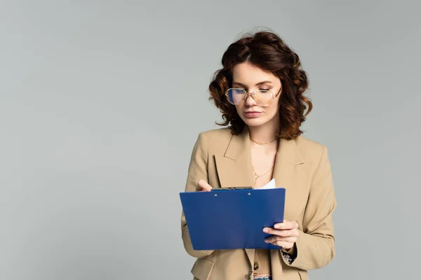 Geschäftsfrau mit Brille und Blazer hält Klemmbrett isoliert auf grau — Stockfoto