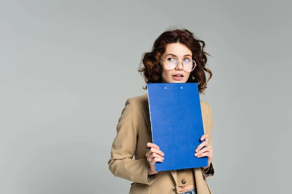 Curious businesswoman in glasses and blazer holding clipboard and looking away isolated on grey — Stock Photo