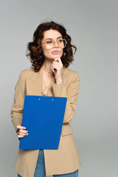 Nachdenkliche Geschäftsfrau mit Brille und Blazer hält Klemmbrett isoliert auf grau — Stockfoto