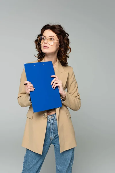 Ricci giovane donna d'affari in occhiali e blazer tenendo appunti isolati su grigio — Foto stock