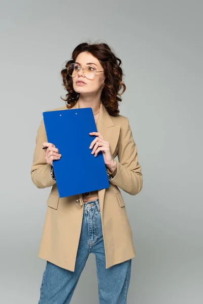 Joven empresaria rizada en gafas y blazer sujetando portapapeles aislado en gris - foto de stock