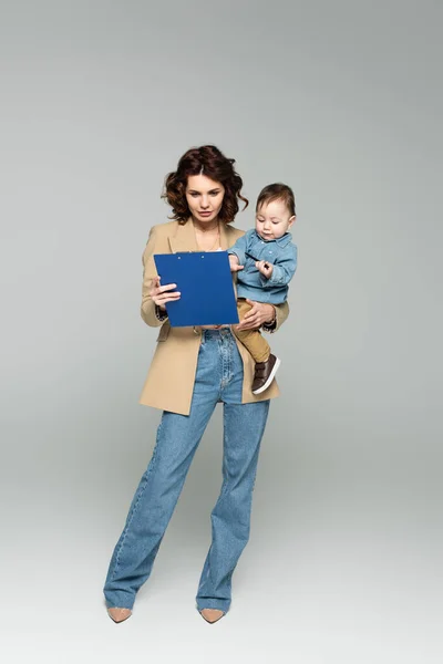 Pleine longueur de femme d'affaires en blazer tenant presse-papiers et bébé garçon dans les bras sur gris — Photo de stock