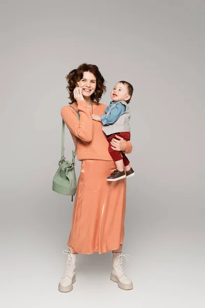 Volle Länge der glücklichen Frau in Satinrock hält in den Armen Kleinkind Sohn und telefoniert auf Handy auf grau — Stockfoto
