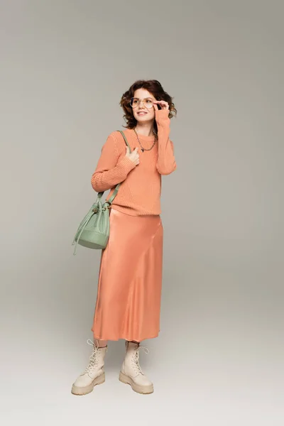 Comprimento total da mulher elegante em suéter e saia de cetim segurando pulseira de couro de saco e ajustando óculos em cinza — Fotografia de Stock