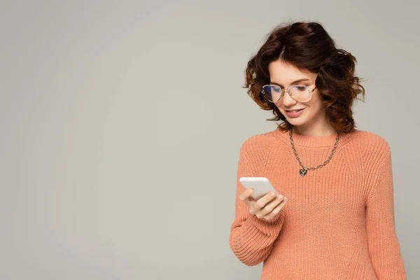 Mujer complacida en gafas de texto en el teléfono inteligente aislado en gris - foto de stock