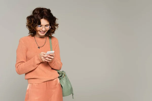Позитивна жінка в окулярах смс на смартфоні ізольована на сірому — стокове фото