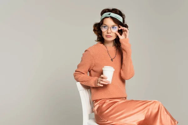 Mujer rizada ajustando gafas y sosteniendo la taza de papel mientras está sentada en la silla aislada en gris - foto de stock