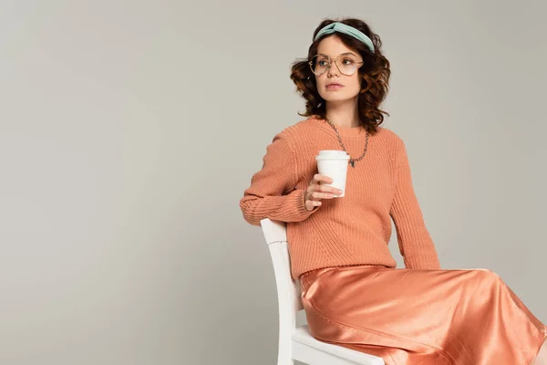 Curly woman in glasses and headband holding paper cup while sitting on chair isolated on grey — Stock Photo