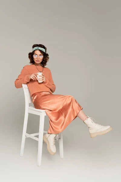 Full length of curly woman in glasses and headband holding paper cup while sitting on chair on grey — Stock Photo