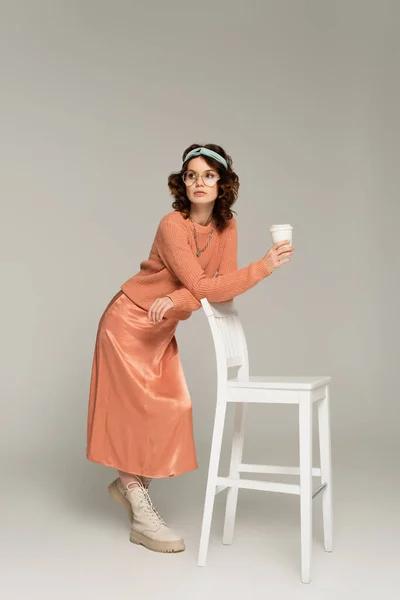 Full length of curly woman in glasses and headband leaning on chair while holding paper cup on grey — Stock Photo