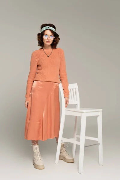 Full length of curly woman in glasses and headband standing near chair on gray — Stock Photo