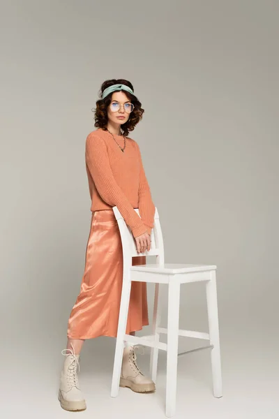 Full length of young woman in glasses and headband standing near chair on gray — Stock Photo