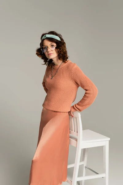 Young woman in glasses and headband holding white chair isolated on gray — Stock Photo