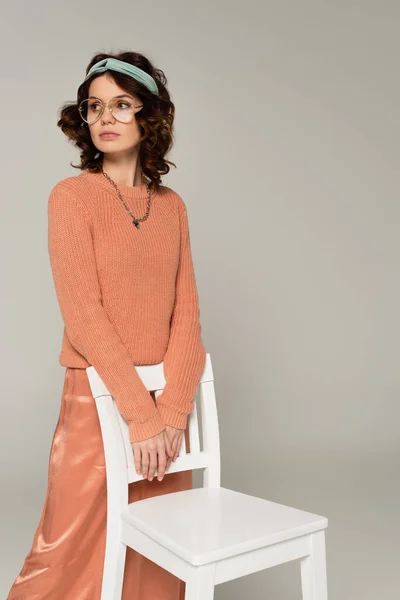 Curly young woman in glasses and headband holding chair isolated on gray — Stock Photo