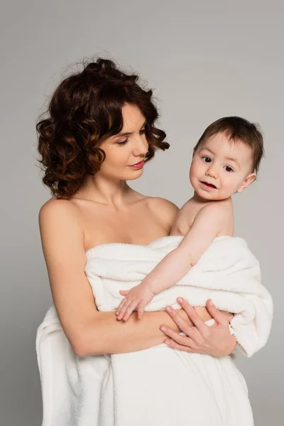 Caring mother with bare shoulders holding son wrapped in towel isolated on grey — Stock Photo