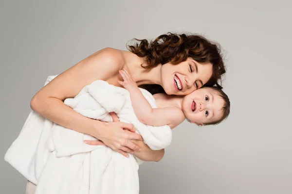 Joyful mother with bare shoulders holding toddler son wrapped in towel isolated on grey — Stock Photo