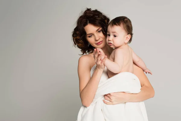 Caring mother with bare shoulders holding hands with baby boy wrapped in towel isolated on grey — Stock Photo