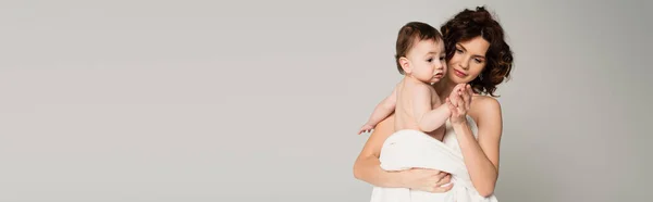 Caring mother with bare shoulders holding hands with baby boy wrapped in towel isolated on grey, banner — Stock Photo