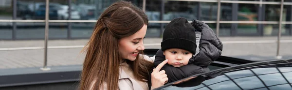 Smiling woman touching cheek of baby boy near car on urban street, banner — Stock Photo