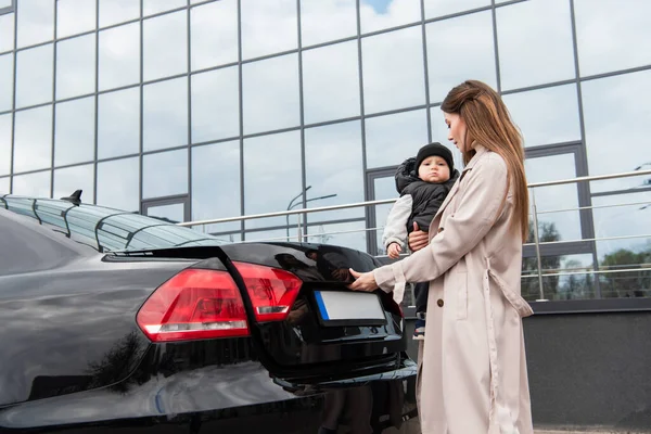 Jovem mulher abertura carro tronco enquanto de pé com criança na rua urbana — Fotografia de Stock