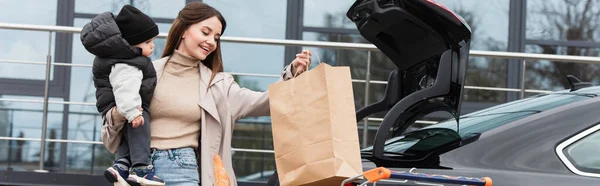 Femme heureuse tenant sac à provisions et fils tout-petit près de la voiture, bannière — Photo de stock