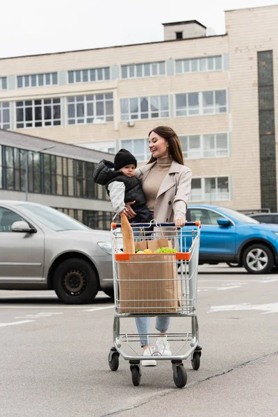 Mujer alegre sosteniendo pequeño hijo mientras camina con carrito de compras - foto de stock