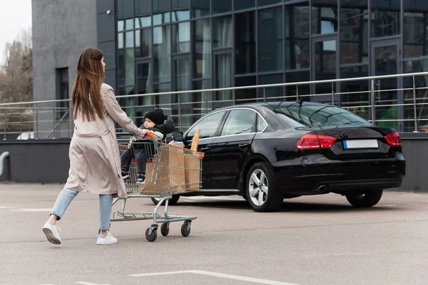 Young mother with purchases and toddler son in shopping cart walking on car parking — Stock Photo