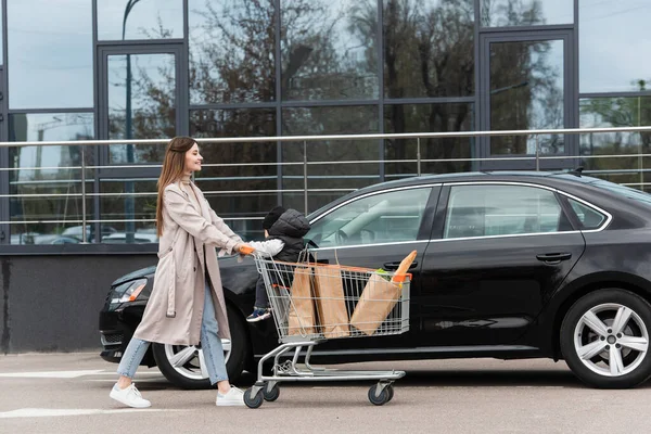 Vista lateral de la mujer joven con bebé niño y bolsas de compras en el carro en el aparcamiento - foto de stock