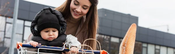 Joven madre sonriendo cerca de niño pequeño hijo sentado en el carro de la compra, pancarta - foto de stock