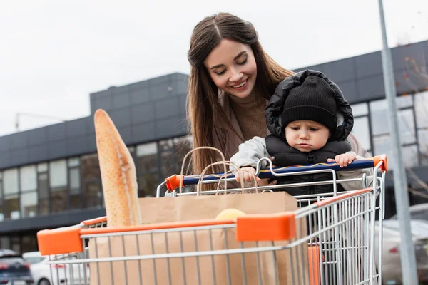 Mujer sonriente con hijo pequeño y compras en carrito de compras al aire libre - foto de stock