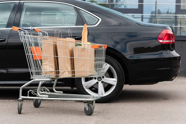 Einkaufstaschen im Einkaufswagen neben schwarzem Auto im Freien — Stockfoto