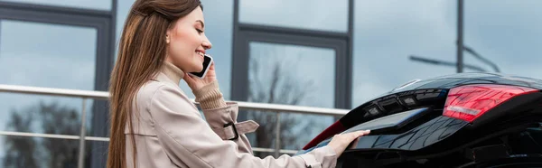 Mujer feliz sonriendo mientras habla en el teléfono móvil cerca del maletero del coche, pancarta - foto de stock