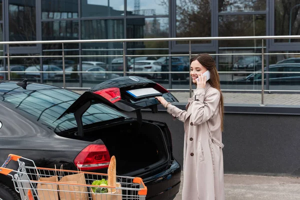 Mujer joven hablando en smartphone cerca de coche y compras en carro - foto de stock